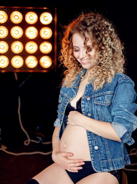 Schwangere junge schöne Mode blonde Modellfrau in Jeansjacke im Studio auf Stuhl sitzend