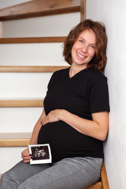 schwangere lächelnde Frau, die auf hölzerner Treppe sitzt und beim Zeigen des Ultraschalls aufwirft