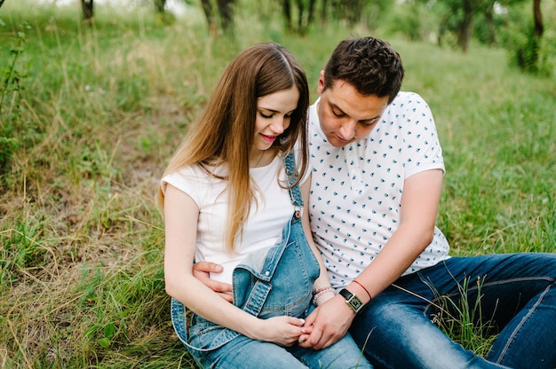 Schwangere Mädchen und ihr Ehemann sind glücklich zu umarmen, Hände auf dem Bauch zu halten und im Freien im Freien auf der Gartenoberfläche zu sitzen