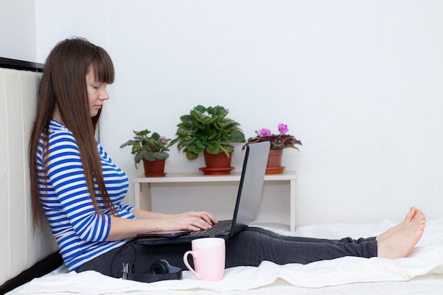 Schwangere mit einem Laptop auf dem Bett. Heimarbeit . Gesundheit & Sicherheit . Mädchen und Fernarbeit. Coronavirus.