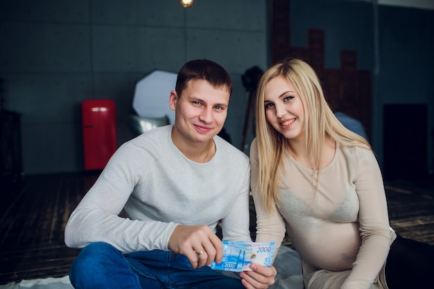 Schwangere mit Geld und Babykleidung sitzen auf einem Sofa des Hauses. Familienbudget.