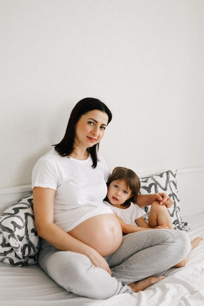 Schwangere Mutter mittleren Alters sitzt zu Hause auf dem Bett und umarmt ihre kleine Tochter