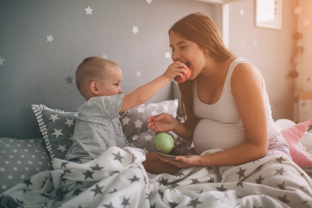 Schwangere mutter und sohn des kleinen jungen essen einen apfel und einen pfirsich im bett, die morgens zu hause sind. lässiger lebensstil im schlafzimmer.