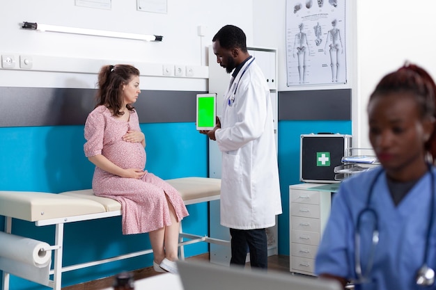 Schwangere Patientin, die im Büro einen vertikalen grünen Bildschirm auf einem Tablet betrachtet. Arzt hält Chroma-Key auf Gerätedisplay für Mockup-Vorlage und isolierten Hintergrund bei Kontrollbesuch.