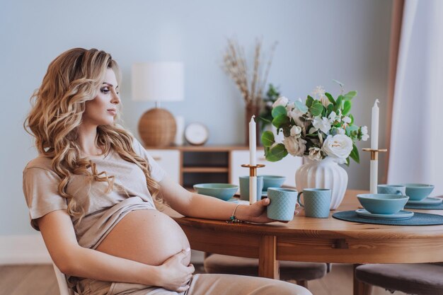 Schwangere Schwedin in beigen T-Shirt-Hosen mit Tasse, die ihren Bauch berührt und am Tisch zu Hause sitzt