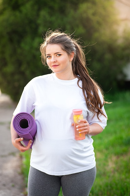 Schwangere steht in der Natur mit Yogamatte und Flasche Wasser. Schwangere vor dem Training