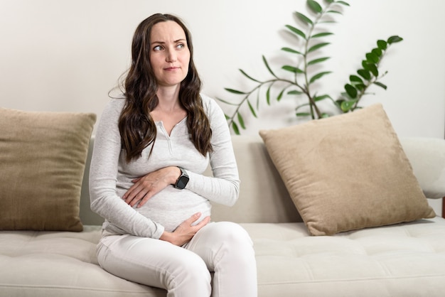 Schwangere verwirrte Frau in grauer Bluse sitzt auf der Couch und denkt, Emotionen, Erwartung