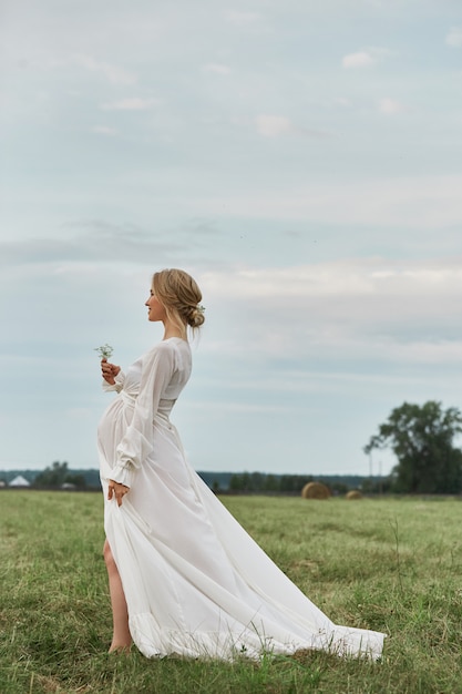 Schwangeres Mädchen geht auf einem Gebiet nahe Heuschober in einem langen weißen Kleid