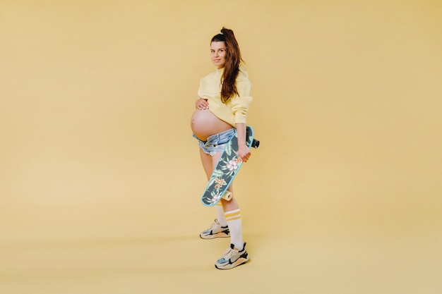 Schwangeres Mädchen in einer gelben Jacke mit einem Skateboard in den Händen auf gelbem Hintergrund