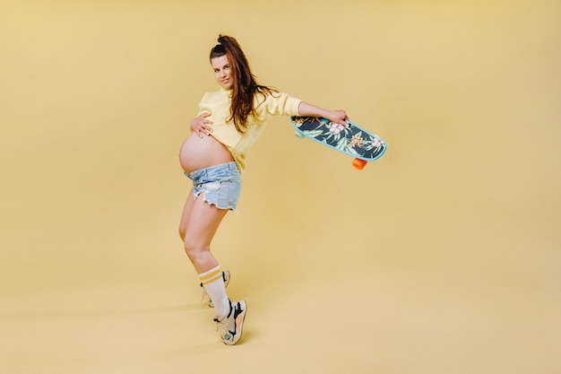 Schwangeres Mädchen in einer gelben Jacke mit einem Skateboard in den Händen auf gelbem Hintergrund.