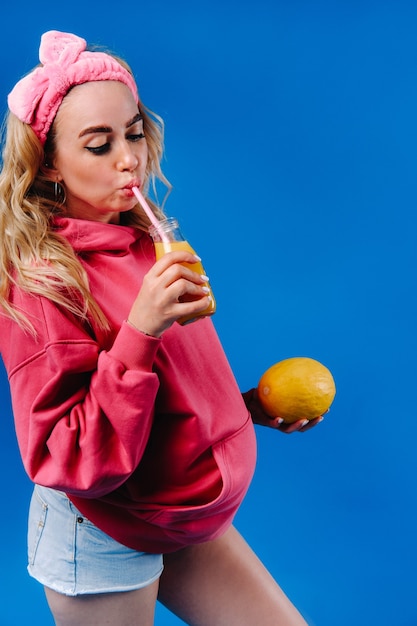 Schwangeres Mädchen in rosa Kleidung mit einer Flasche Saft und einer Melone auf blauem Hintergrund.