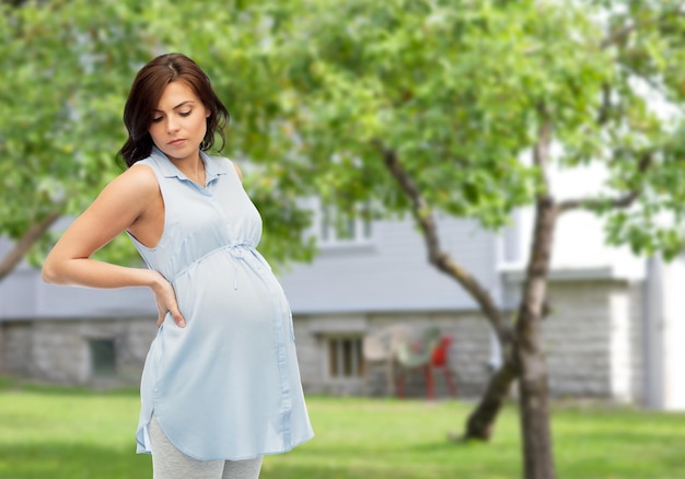 schwangerschaft, gesundheit, menschen und erwartungskonzept - schwangere frau, die ihren rücken berührt und unter rückenschmerzen über sommergarten und haushintergrund leidet
