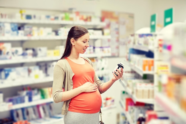 schwangerschaft, medizin, pharmazie, gesundheitswesen und personenkonzept - glückliche schwangere frau liest etikett auf medikamentenglas in der apotheke