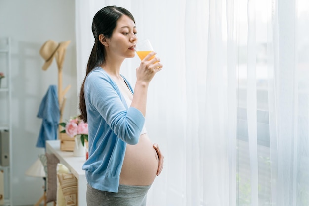 schwangerschaft menschen gesundes essen konzept. glücklich lächelnde elegante schwangere frau, die zu hause saft-smoothie am fenster im wohnzimmer trinkt. charmante Mutter des jungen Mädchens genießt Elternschaft mit frischen Lebensmitteln und Getränken