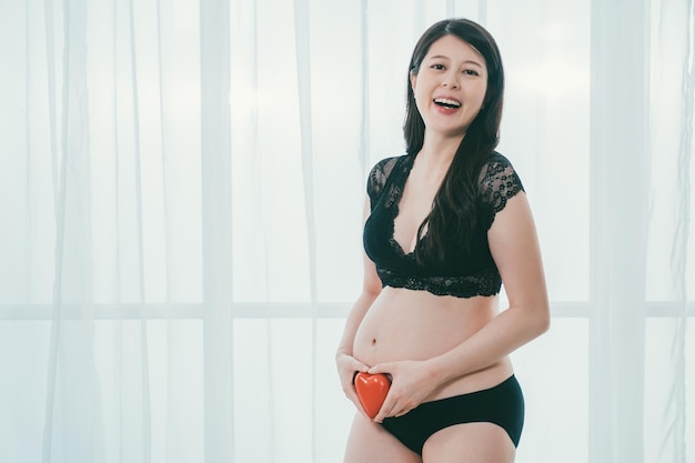 Schwangerschaft Mutterschaft. Fröhliche asiatische schwangere Mutter, die rote Herzform am nackten Bauch hält und fröhlich lacht. Fashion Future Mom in schwarzem Spitzen-BH und Höschen zeigt Liebe zu ungeborenen Zwillingen Baby