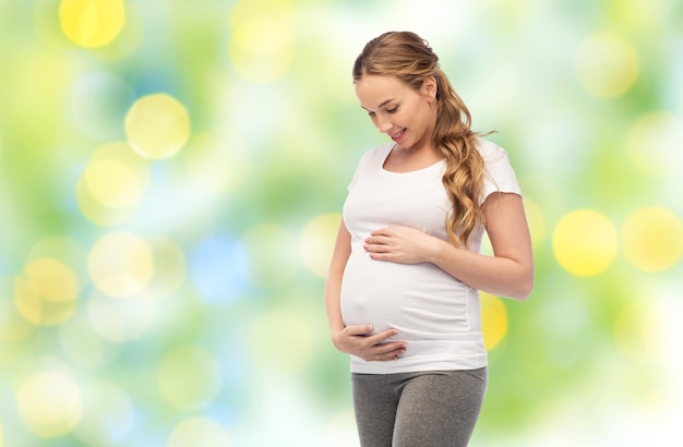 Schwangerschaft, Mutterschaft, Menschen und Erwartungskonzept - glückliche schwangere Frau berührt ihren dicken Bauch vor dem Hintergrund grüner Sommerlichter