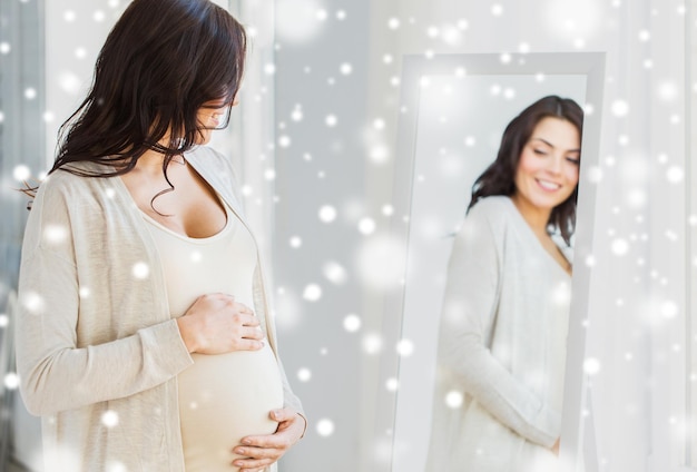Schwangerschaft, Mutterschaft, Winter, Menschen und Erwartungskonzept – Nahaufnahme einer glücklichen schwangeren Frau, die sich zu Hause über dem Schnee spiegeln möchte
