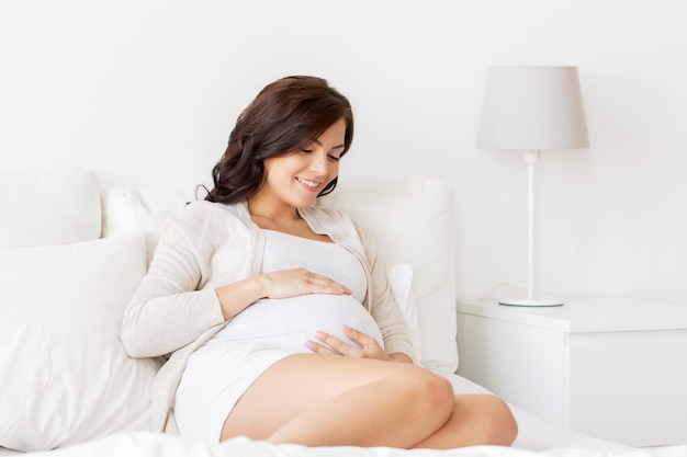 schwangerschaft, ruhe, menschen und erwartungskonzept - glückliche schwangere frau, die auf dem bett liegt und zu hause ihren bauch berührt