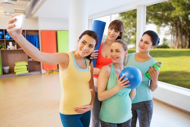 Foto schwangerschaft, sport, fitness, menschen und gesundes lebensstilkonzept - gruppe glücklicher schwangerer frauen mit sportsachen, die selfie per smartphone im fitnessstudio über natürlichem fensteransichtshintergrund machen