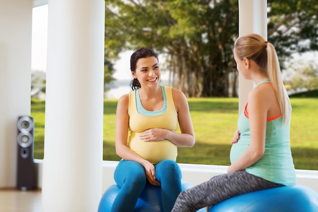 schwangerschaft, sport, fitness, menschen und gesundes lebensstilkonzept - zwei glückliche schwangere frauen sitzen und reden auf bällen im fitnessstudio über natürlichem fensteransichtshintergrund