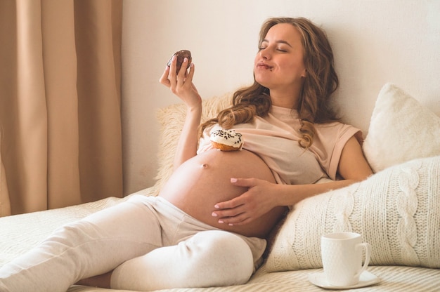 Schwangerschaft und Ernährung. Schwangere Frau genießt Donuts und Tee im Bett, freier Raum