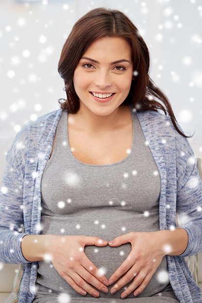 Schwangerschaft, Winter, Weihnachten, Menschen und Erwartungskonzept – glückliche schwangere Frau macht zu Hause über Schnee eine Herzgeste