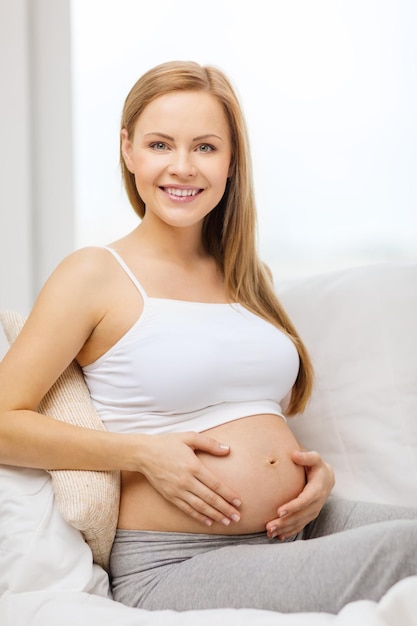 Schwangerschafts-, Mutterschafts- und Glückskonzept - glückliche schwangere Frau, die auf dem Sofa sitzt und ihren Bauch berührt