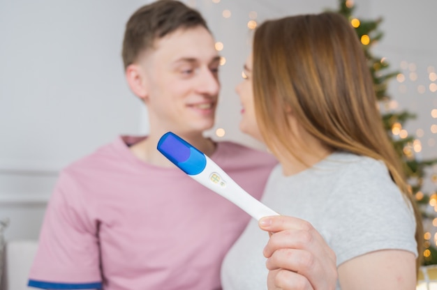 Schwangerschaftstest in der Hand auf dem Hintergrund eines defokussierten lächelnden jungen Paares zu Hause, Weihnachtszeit.