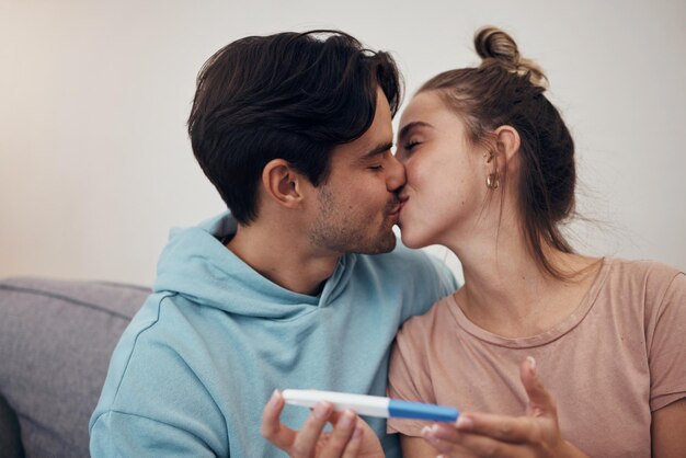 Schwangerschaftstest und Pärchenkuss im heimischen Wohnzimmer für gute Nachrichten oder positive Ergebnisse. Romantischer Mann und schwangere Frau mit Stock auf dem Sofa freuen sich gemeinsam über den Erfolg einer zukünftigen Mutterschaft oder IVF-Fruchtbarkeit