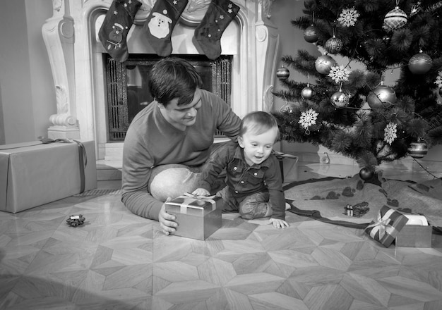 Schwarz-Weiß-Bild des jungen Vaters, der mit seinem 1-jährigen Jungen auf dem Boden neben dem Weihnachtsbaum spielt