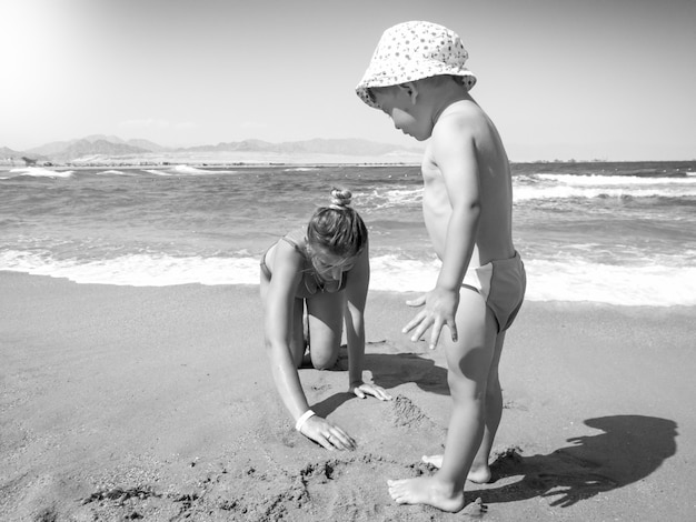 Schwarz-Weiß-Bild des kleinen Kleinkindjungen, der mit junger Mutter am Meeresstrand spielt. Familie, die sich während der Sommerferien entspannt und gute Zeit hat.