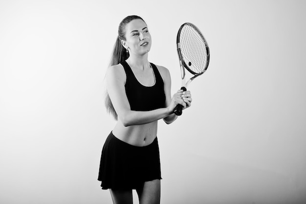 Schwarz-Weiß-Porträt einer schönen jungen Spielerin in Sportkleidung mit Tennisschläger im Stehen vor weißem Hintergrund