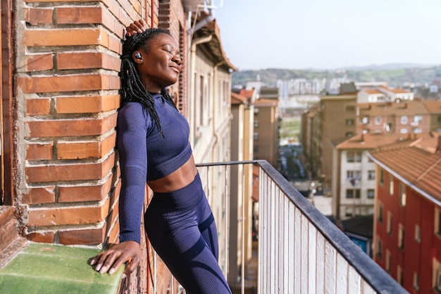 Schwarze Afro-Frau in Sportkleidung, die Musik über Kopfhörer hört, sehr glücklich auf dem Balkon ihres Hauses, weil sie aufgrund der Covid19-Coronavirus-Pandemie zu Hause mit dem Training beginnen wird