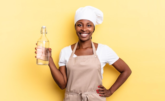Schwarze Afro-Kochfrau, die glücklich mit einer Hand auf der Hüfte und selbstbewusster, positiver, stolzer und freundlicher Haltung lächelt, die eine Wasserflasche hält