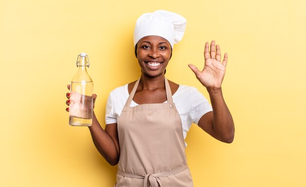 Schwarze Afro-Kochfrau, die lächelt und freundlich aussieht, die Nummer fünf oder fünf mit der Hand nach vorne zeigt, herunterzählt und eine Wasserflasche hält