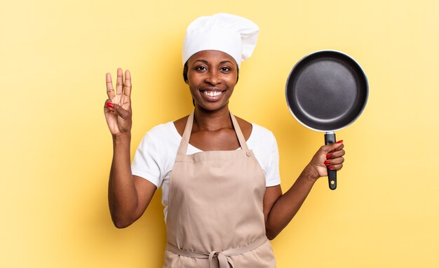 Schwarze afro-kochfrau, die lächelt und freundlich aussieht, nummer drei oder dritte mit der hand nach vorne zeigt, herunterzählt