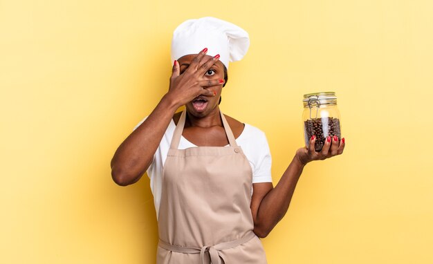 Schwarze Afro-Kochfrau, die schockiert, verängstigt oder verängstigt aussieht, das Gesicht mit der Hand bedeckt und zwischen den Fingern späht. Kaffeebohnen-Konzept