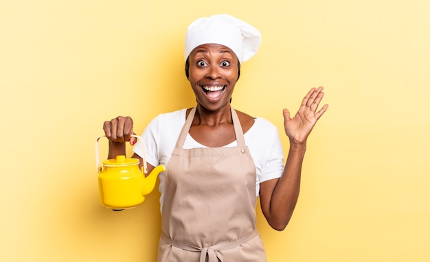 Schwarze Afro-Kochfrau, die sich glücklich, aufgeregt, überrascht oder schockiert fühlt, lächelt und erstaunt über etwas Unglaubliches. Teekannenkonzept