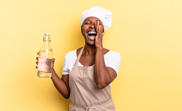 Schwarze Afro-Kochfrau, die sich glücklich, aufgeregt und positiv fühlt, mit den Händen neben dem Mund einen großen Schrei ausstößt und eine Wasserflasche hält