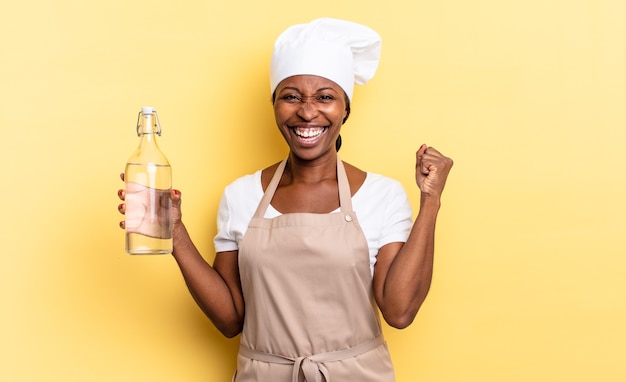 Schwarze Afro-Kochfrau, die sich schockiert, aufgeregt und glücklich fühlt, lacht und den Erfolg feiert und sagt wow! eine Wasserflasche halten