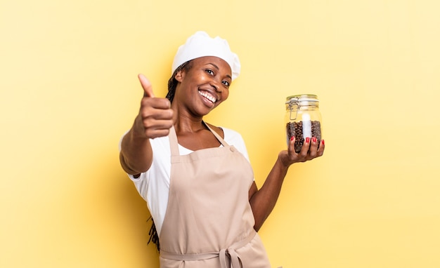 Schwarze Afro-Kochfrau, die sich stolz, sorglos, selbstbewusst und glücklich fühlt und positiv mit Daumen nach oben lächelt. Kaffeebohnen-Konzept