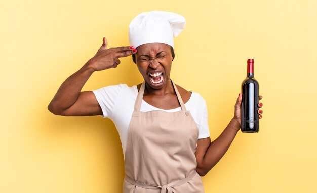 Schwarze Afro-Kochfrau, die unglücklich und gestresst aussieht, Selbstmordgeste, die Waffenzeichen mit der Hand macht und auf den Kopf zeigt. Weinflaschenkonzept