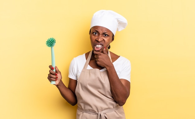 Schwarze Afro-Kochfrau mit weit geöffnetem Mund und Augen und Hand am Kinn, die sich unangenehm schockiert fühlt und was oder wow sagt. Reinigung Geschirr Konzept