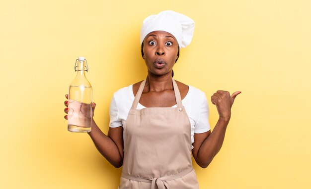 Schwarze Afro-Köchin, die erstaunt und ungläubig auf ein Objekt an der Seite zeigt und sagt: Wow, unglaublich, hält eine Wasserflasche