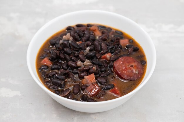 Schwarze Bohnen mit Fleisch und Würstchen in weißer Schüssel auf Keramik