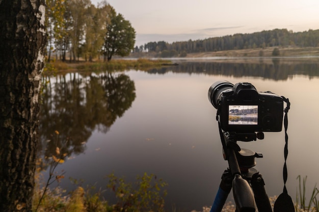 Schwarze Digitalkamera auf Stativ, die neblige Landschaft am frühen Morgen am Herbstsee mit selektivem Fokus aufnimmt