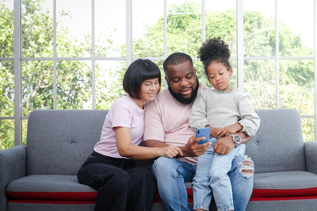 Schwarze Familieneltern und Tochter sitzen auf dem Sofa im Wohnzimmer Sie führen Videoanrufe online auf Smartphones durch. Genießen Sie das Leben zu Hause. Familienkonzept, Einsatz von Technologie zur Kommunikation.