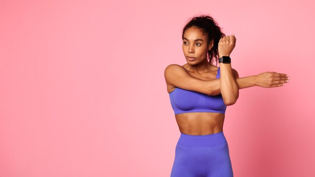 Schwarze Fitnessdame trainiert und streckt die Arme vor rosa Hintergrund