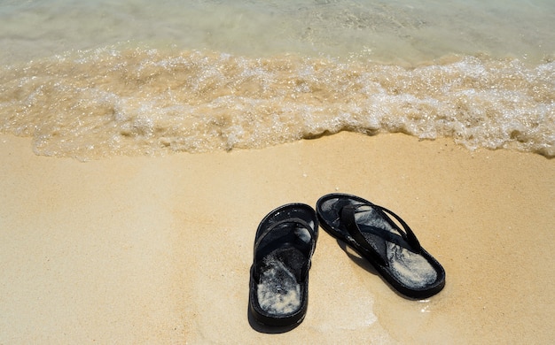 Schwarze Flipflopsandale auf Sandstrand und Seewelle Sommerfeiertag sonnig mein freier Tag concep