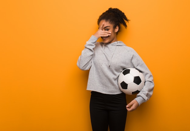 Schwarze Frau der jungen Eignung verlegen und gleichzeitig lachend. Einen Fußball halten.